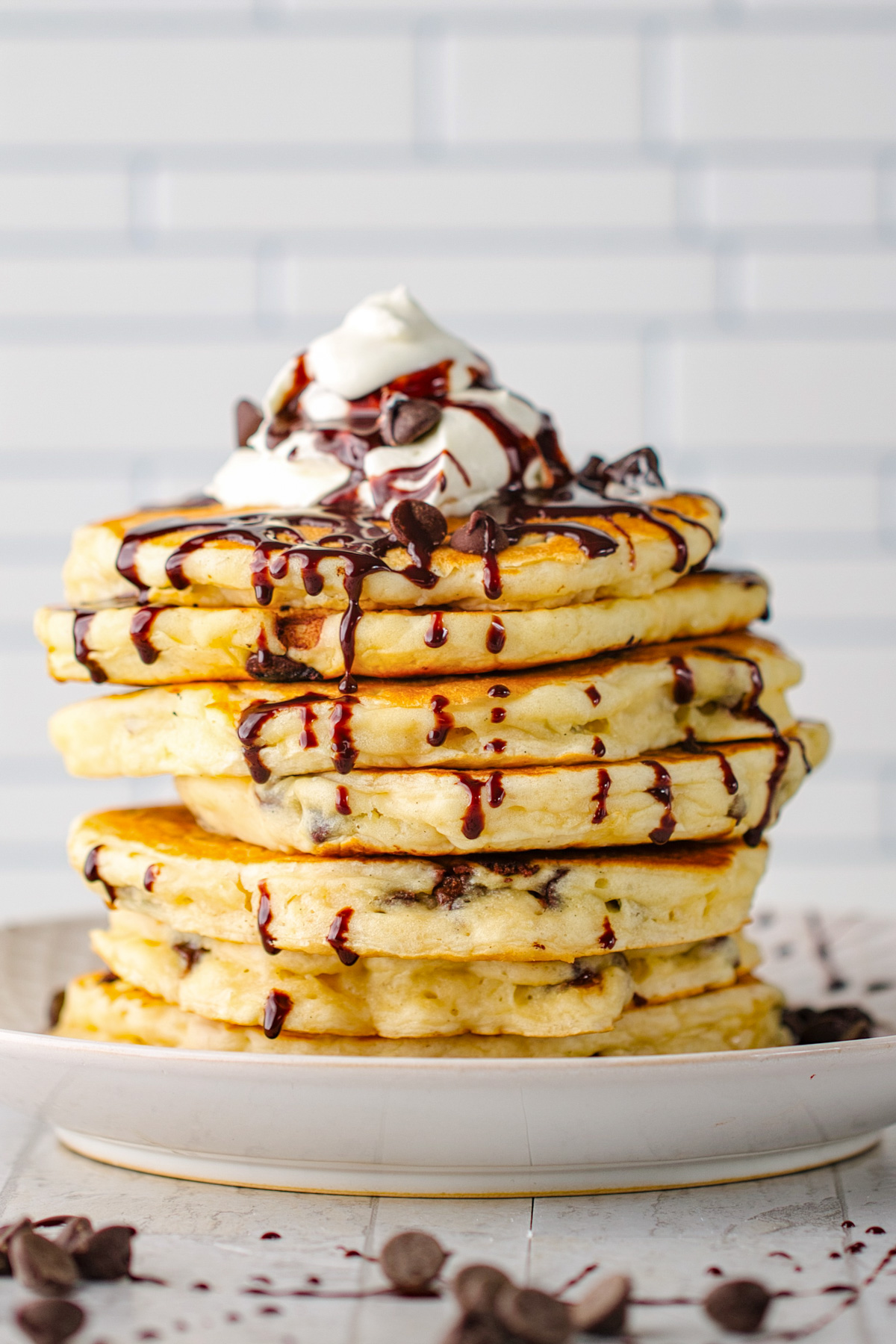 A stock of chocolate chip pancakes topped with whipped cream, with chocolate sauce drizzled over the top and down the sides of the pancakes.