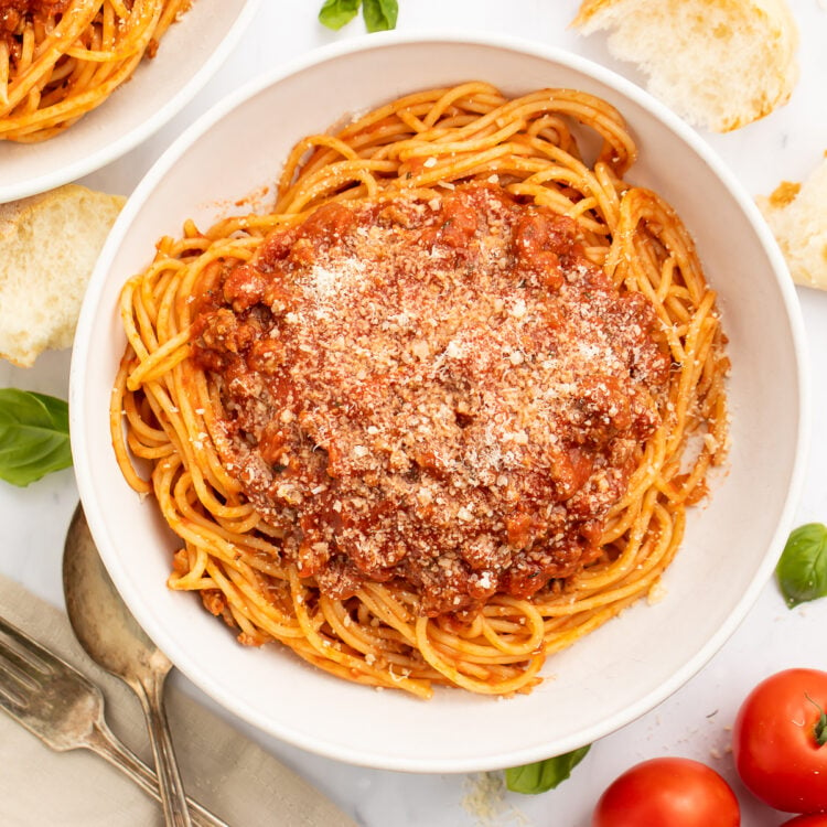 Overhead view of spaghetti noodles coated in rich, red Instant Pot spaghetti sauce in a large white pasta bowl on a table with tomatoes and silverware.