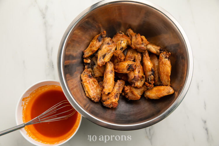 Cooked Whole30 buffalo wings in a large silver mixing bowl, next to Whole30 buffalo sauce in a small white bowl.