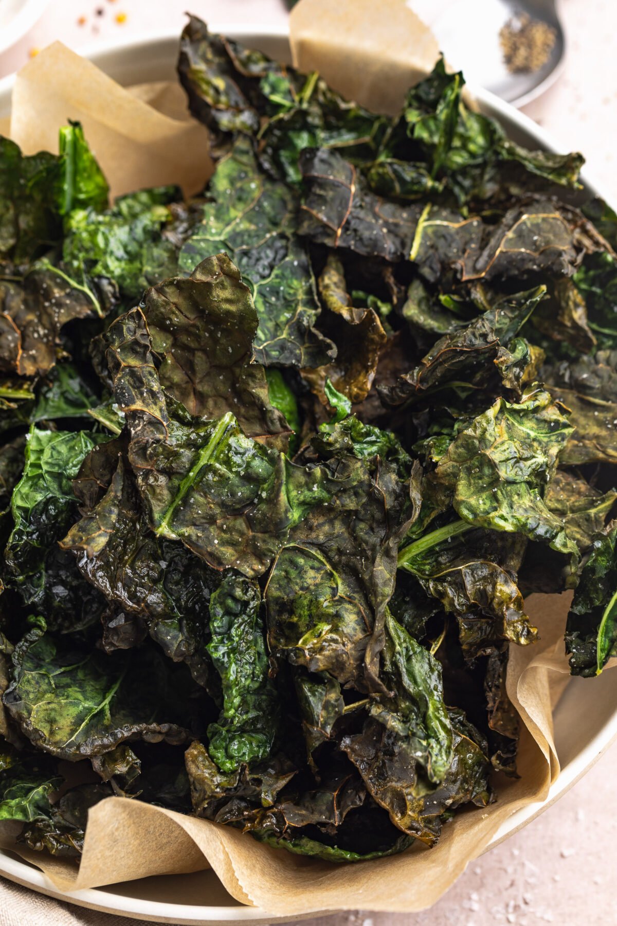 A large bowl, lined with a sheet of tan parchment paper, filled with crispy crunchy deep green kale chips cooked in the air fryer.