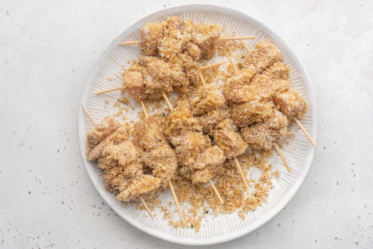 Breaded boneless, skinless chicken breast pieces threaded onto wooden skewers and stacked on a white plate.