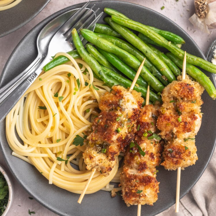Top down view of three chicken spiedini skewers plated on a dark grey plate next to a small portion of angel hair pasta and a pile of bright green beans.