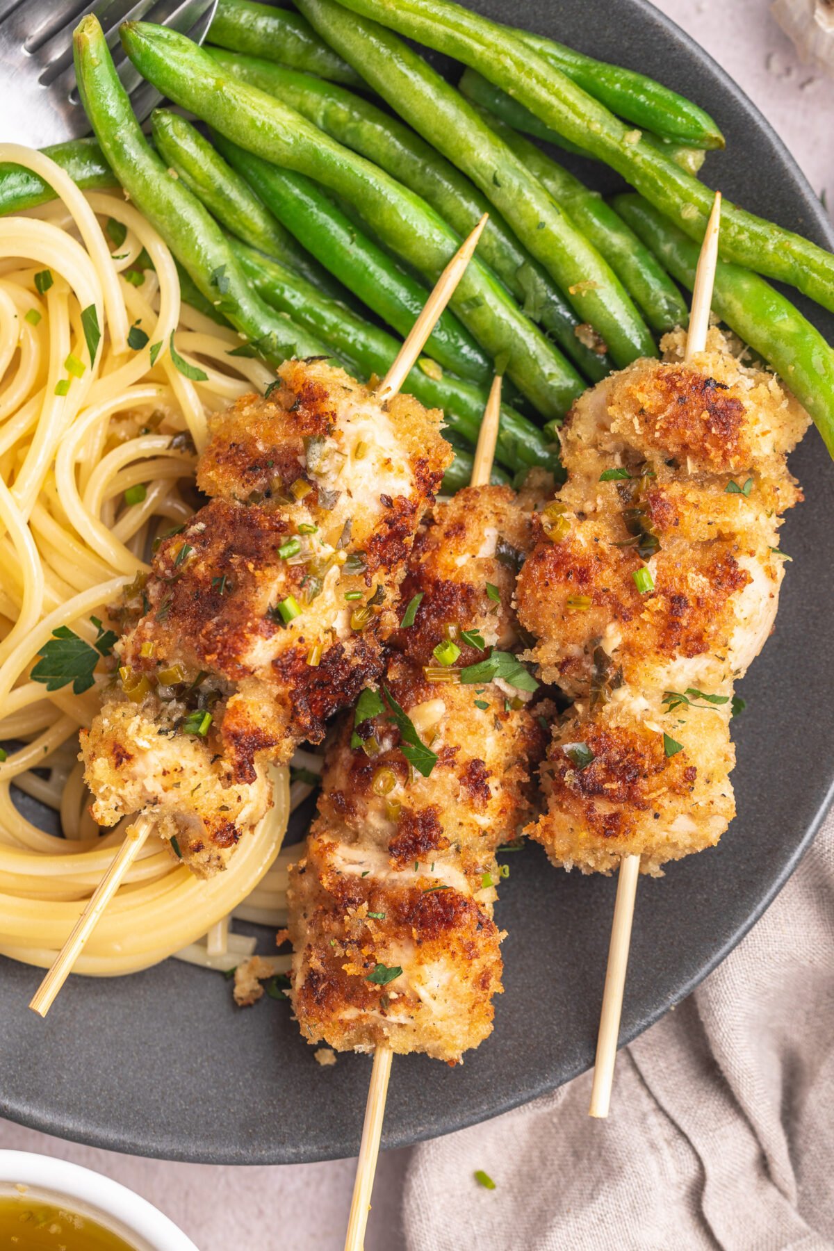 Three chicken spiedini skewers on a dark grey plate with angel hair pasta and bright green beans topped with an herb butter sauce.