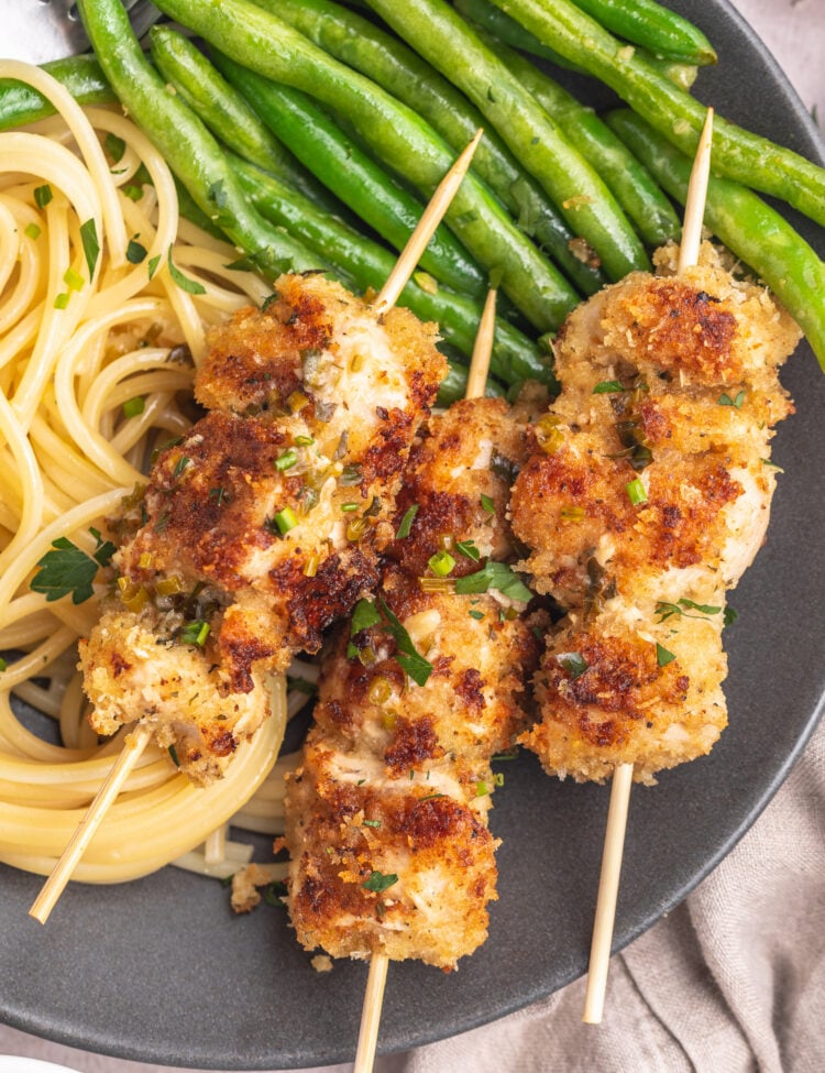 Three chicken spiedini skewers on a dark grey plate with angel hair pasta and bright green beans topped with an herb butter sauce.