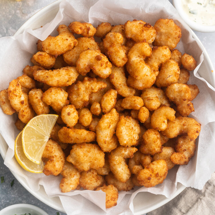 A large white bowl filled to the brim with golden, crispy popcorn shrimp cooked in the air fryer, with a lemon wedge to garnish and ramekins of dipping sauce surrounding.