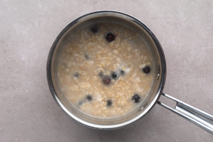 Cooked oatmeal with some water and blueberries in a silver saucepan with a single handle.