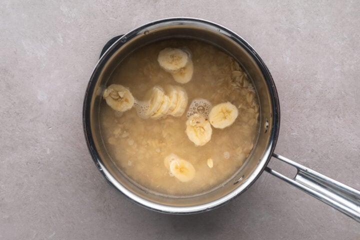 Top-down look at a silver saucepan containing uncooked rolled oats under water, with banana slices floating on top.
