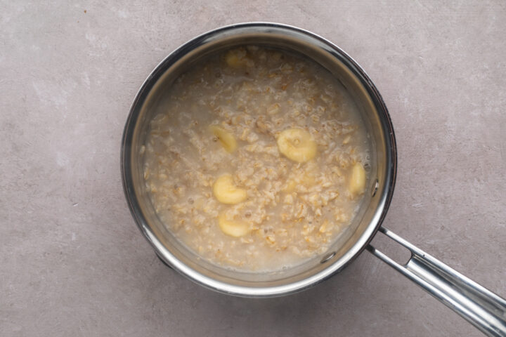 Cooked rolled oats with slices of banana mixed throughout in a silver saucepan with a single handle.