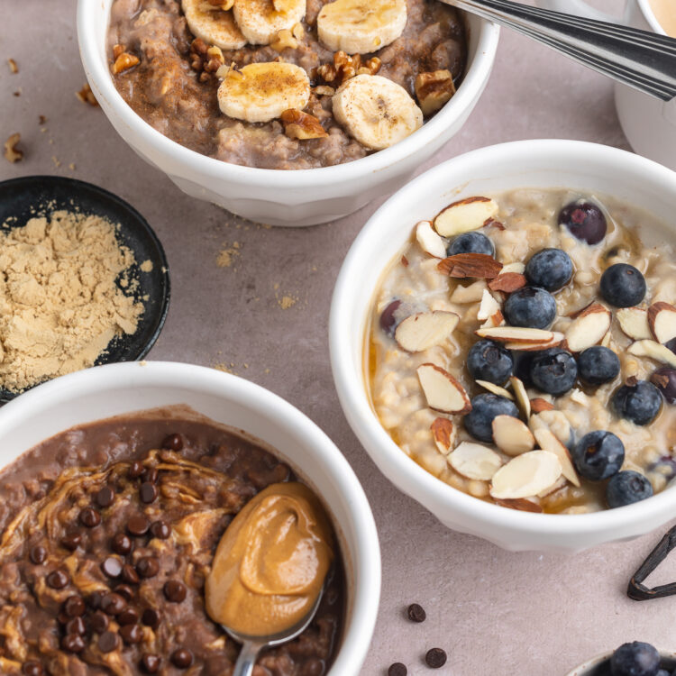 Overhead angled view of 3 different flavors of protein oatmeal, each in a small white ramekin on a neutral tabletop.
