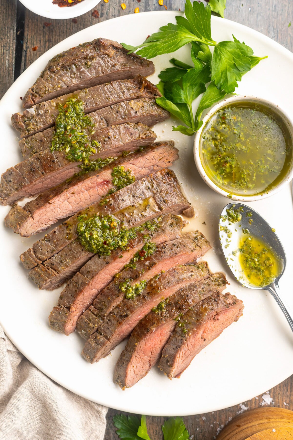 Overhead view of a sous vide flank steak, sliced thin and arranged on a white platter and topped with fresh green chimichurri sauce, next to a ramekin of chimichurri.