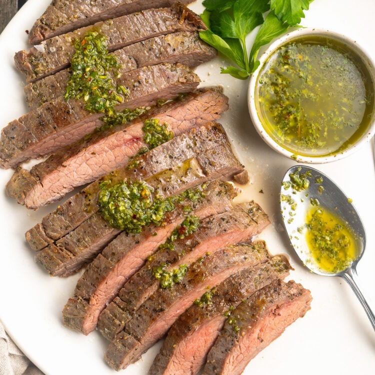 Overhead view of a sous vide flank steak, sliced thin and arranged on a white platter and topped with fresh green chimichurri sauce, next to a ramekin of chimichurri.
