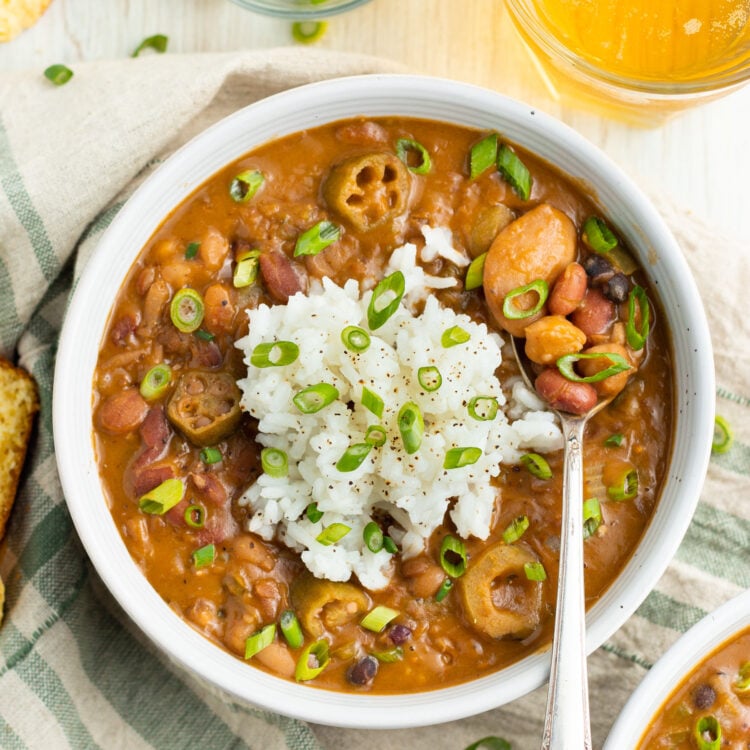 Overhead photo of a white bowl filled with reddish-brown Cajun 15-bean vegetarian gumbo, topped with rice and slices of green onion.