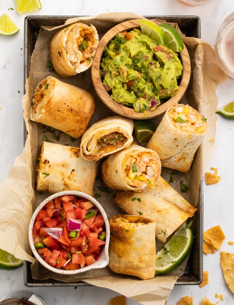 Top-down view of frozen burritos cooked in the area fryer, halved and arranged in a baking pan with guacamole and fresh salsa.