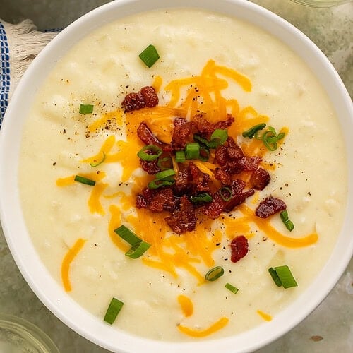 Creamy old-fashioned potato soup in a white bowl topped with shredded cheddar cheese, bacon, and green onions.