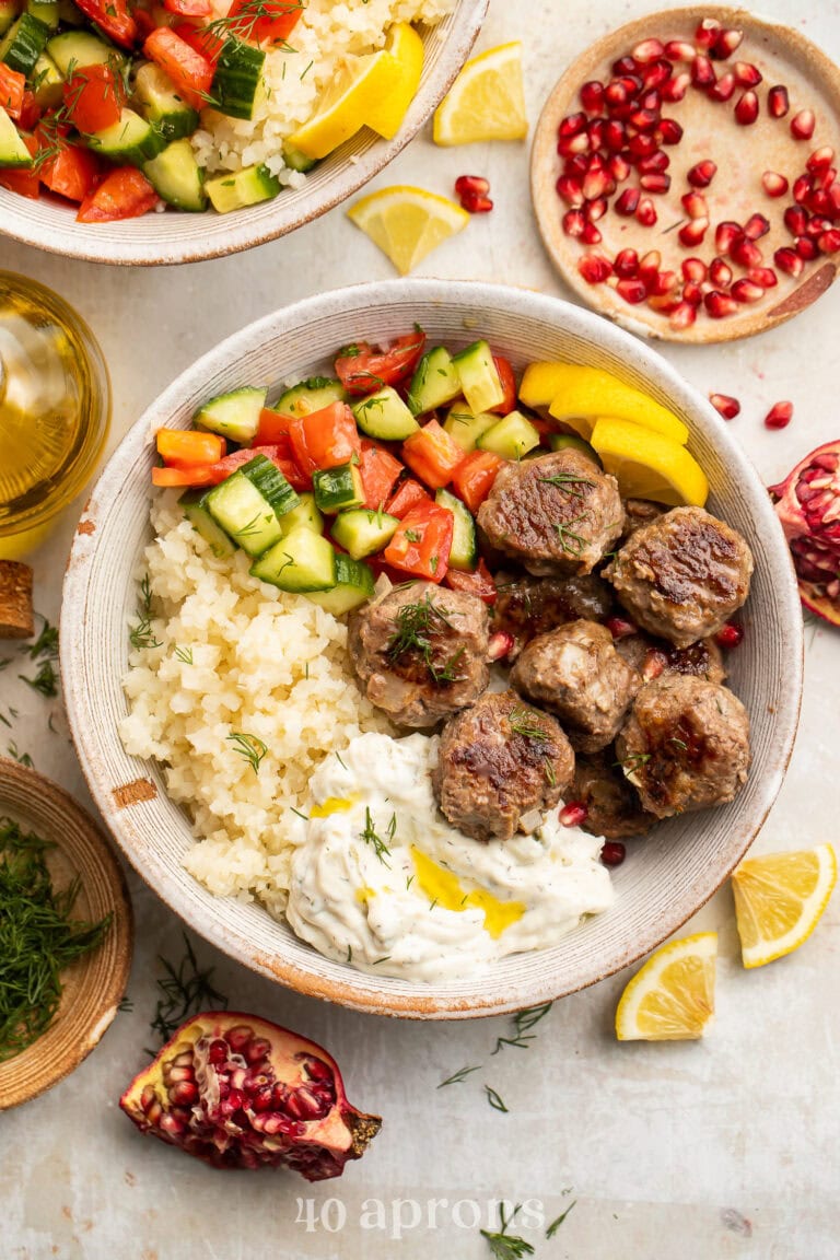 Whole30 gyro meatballs in a bowl with cucumber salsa, cauliflower rice, and Whole30 tzatziki on a table surrounded by scattered ingredients.