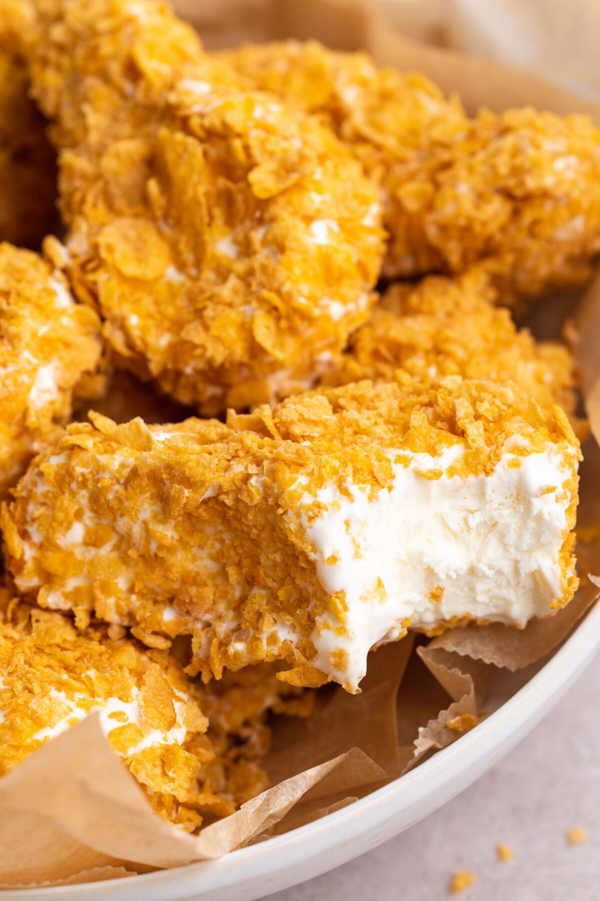 Fried chicken ice cream drumsticks coated in white chocolate and crushed cornflakes. A bite is missing from the forward-most drumstick, showing vanilla ice cream inside.