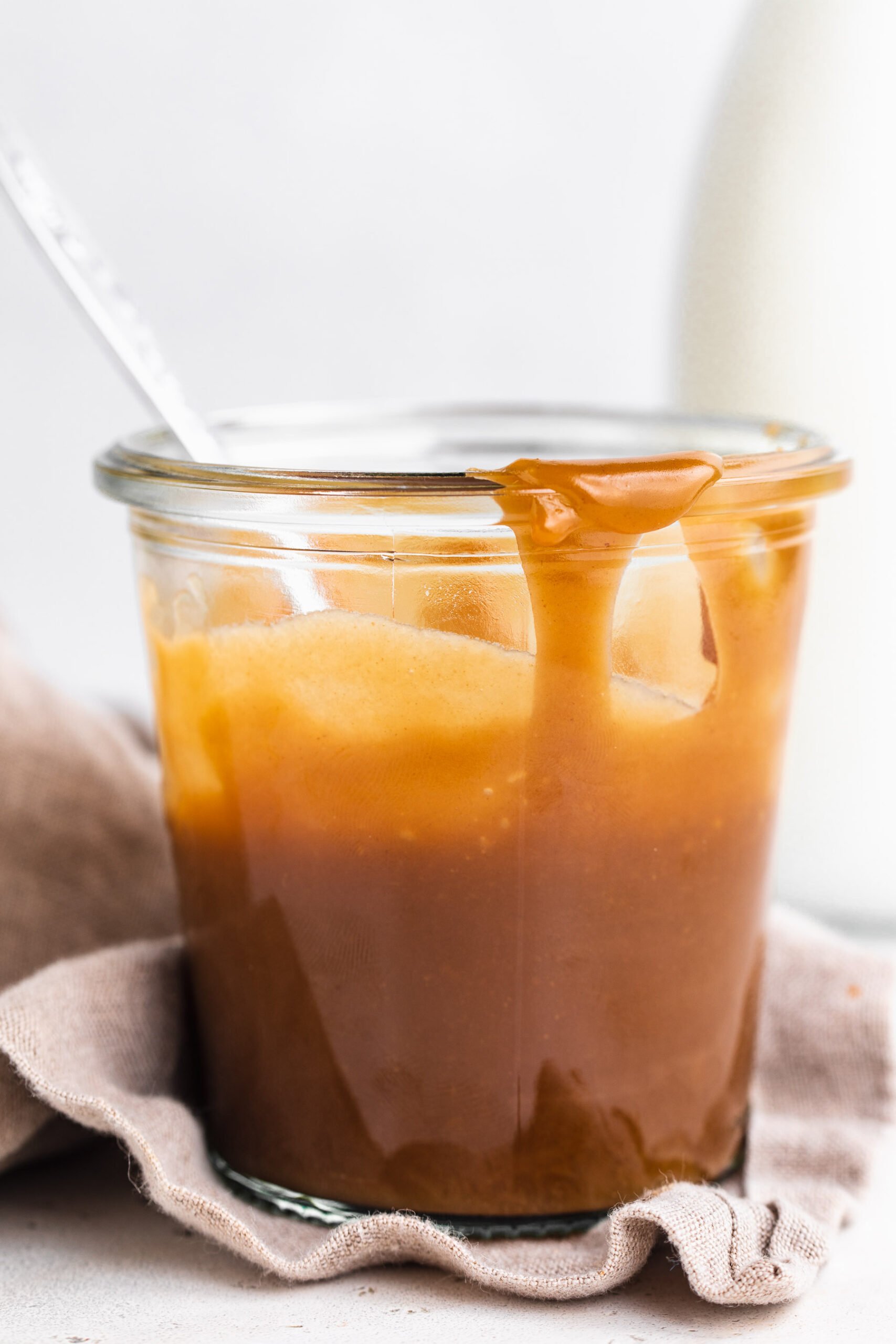 Side view of a glass jar of keto caramel sauce in front of a white background with a neutral napkin around the jar base.