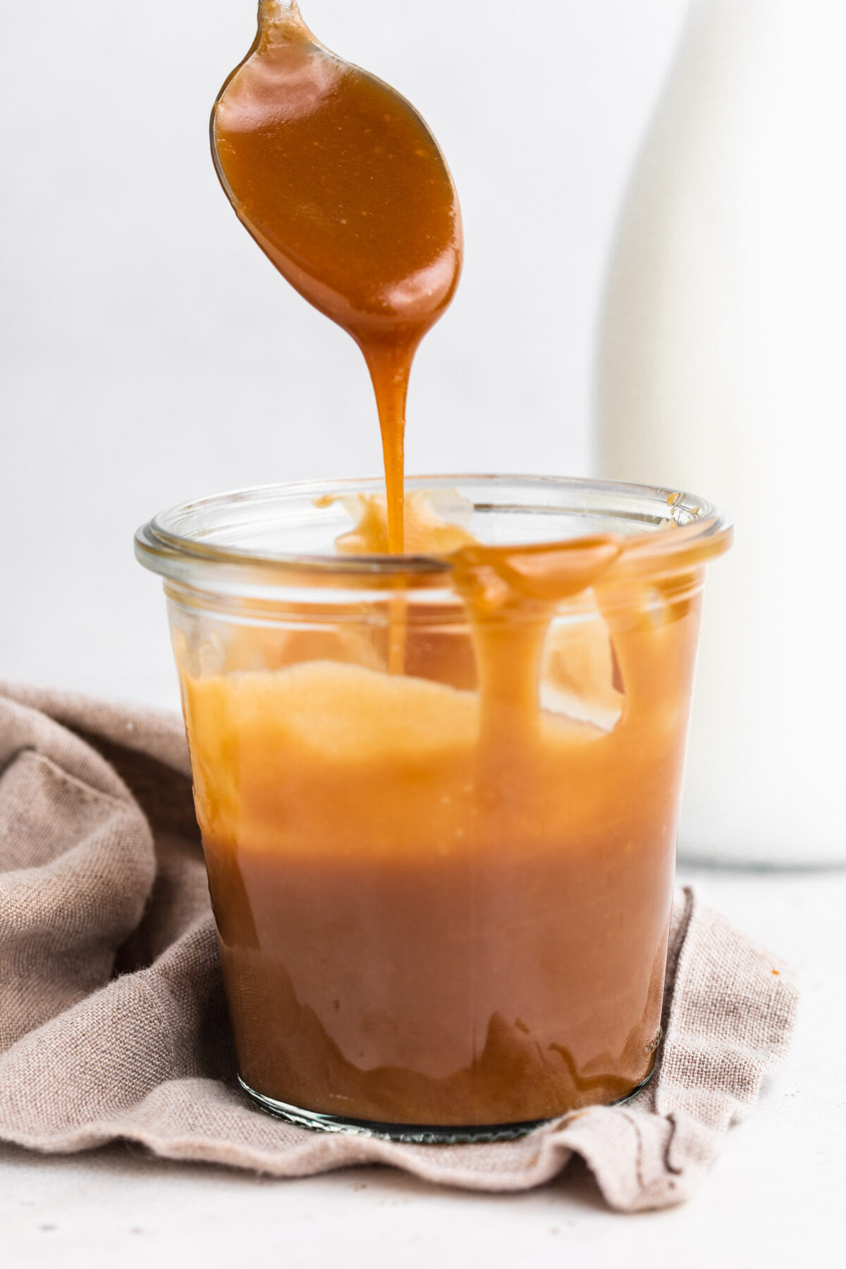 Side view of a jar of keto caramel sauce. A silver spoon hovers above the glass jar, drizzling caramel back in to the jar.