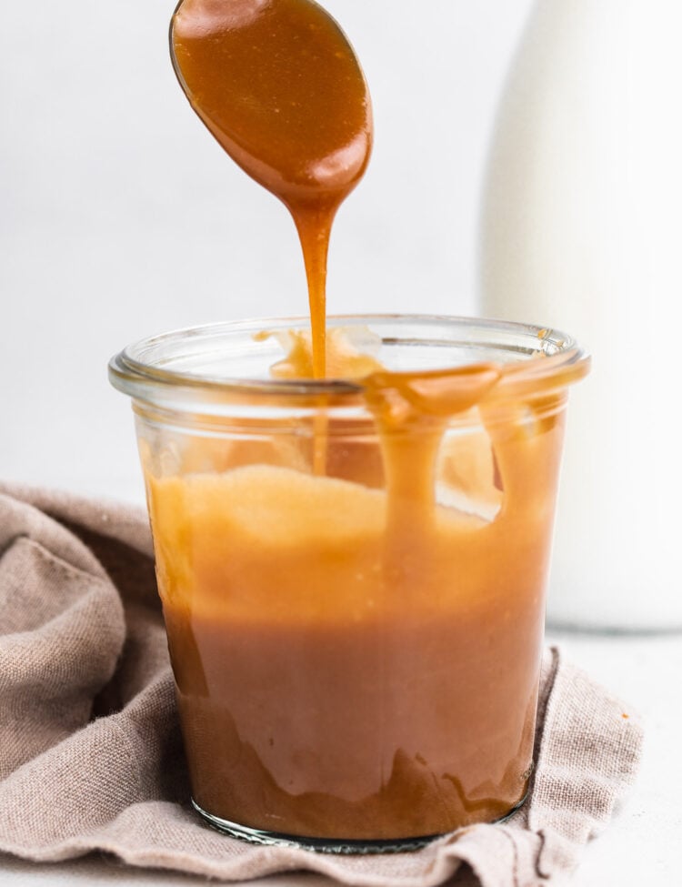 Side view of a jar of keto caramel sauce. A silver spoon hovers above the glass jar, drizzling caramel back in to the jar.