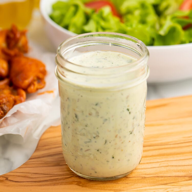 A mason jar of ranch dressing peppered with green specks from fresh herbs sits on a wooden cutting board, in front of a bright green salad and orange chicken wings.