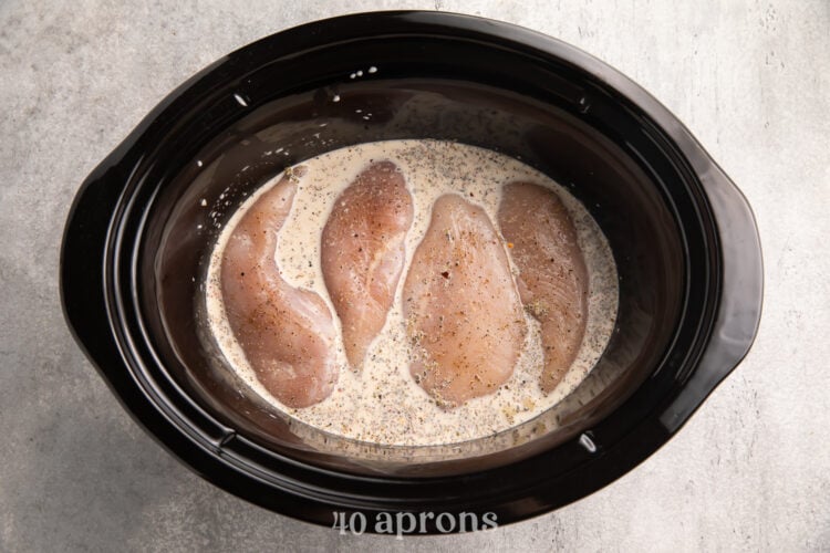Top-down view of 4 chicken breasts in a Crockpot, surrounded by a creamy mixture of heavy cream and various spices.