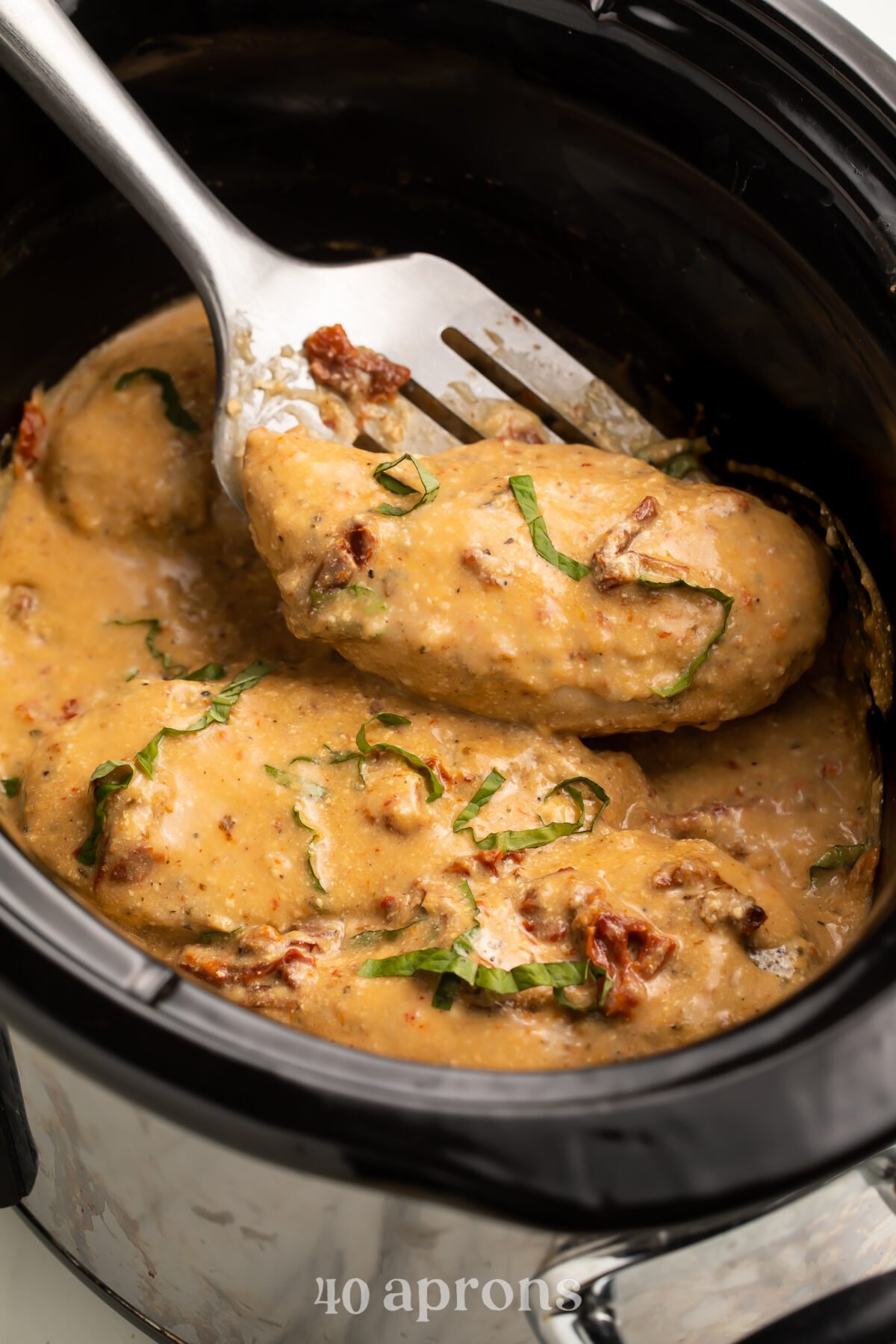 A Marry Me Chicken breast being lifted out of a Crockpot of sauce with a silver spatula.