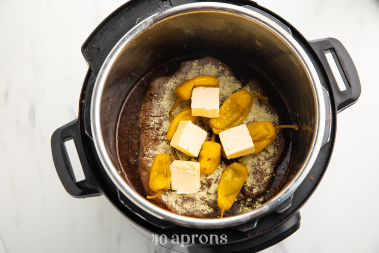 Top-down view of water, chuck roast, seasoning, pepperoncini, and butter in an Instant Pot.