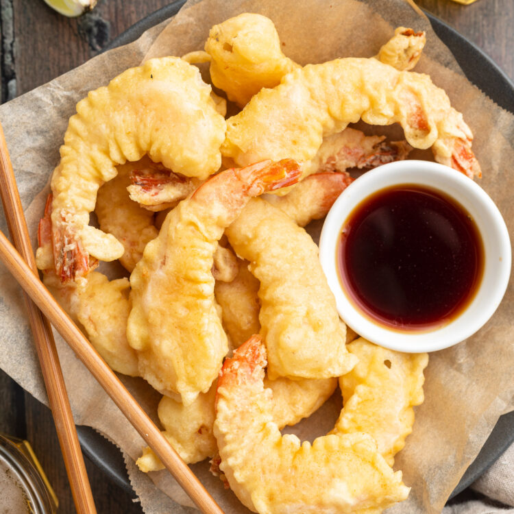 Zoomed-out, overhead photo of shrimp tempura on parchment paper next to a bowl of tempura sauce and chopsticks.
