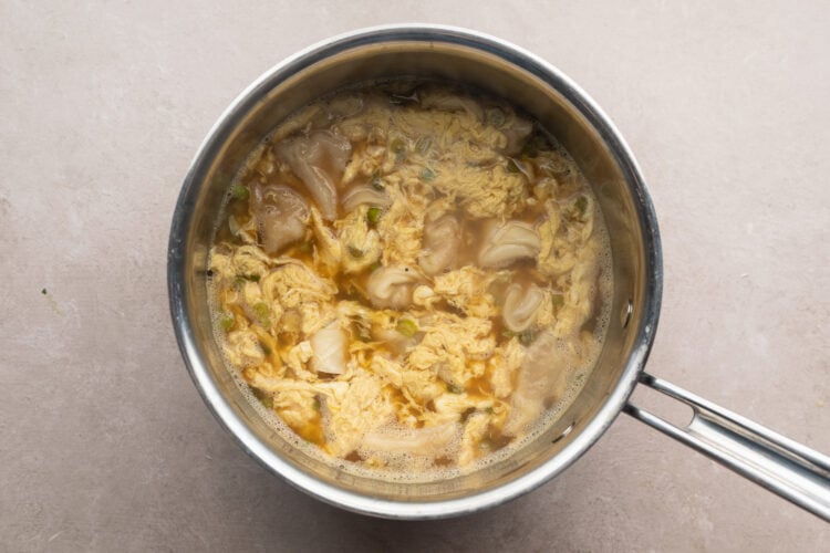 Wontons added to wonton egg drop soup with scrambled egg ribbons in silver saucepan.