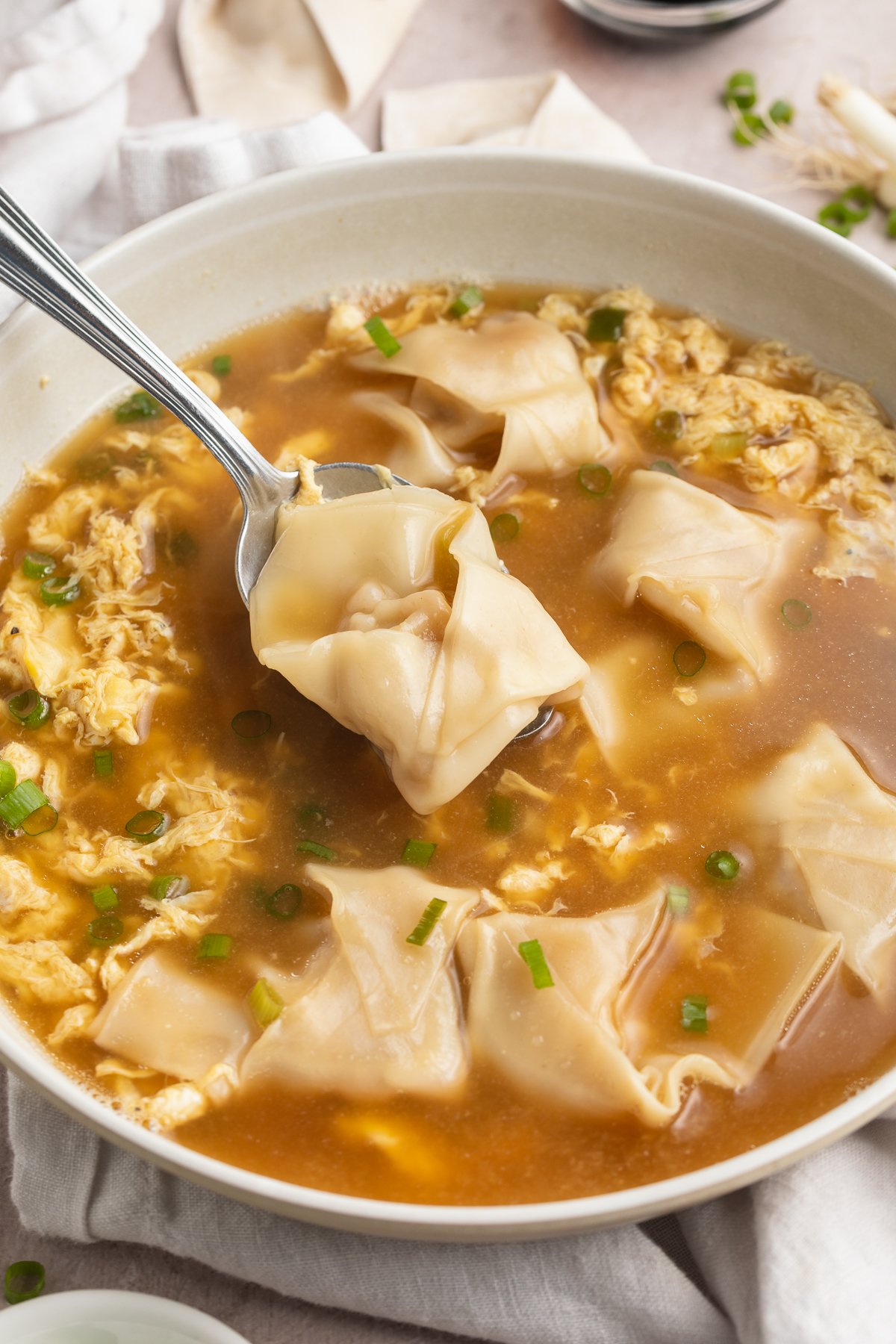 Angled photo of a bowl of wonton egg drop soup. A spoon lifts a wonton up and out of the broth.