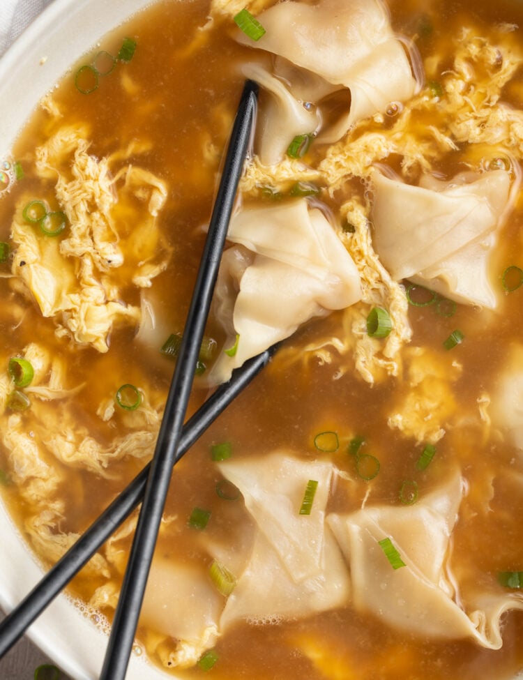 Overhead view of a bowl of wonton egg drop soup. 2 black chopsticks rest in the bowl of soup, and the right half of the bowl is out of the frame.