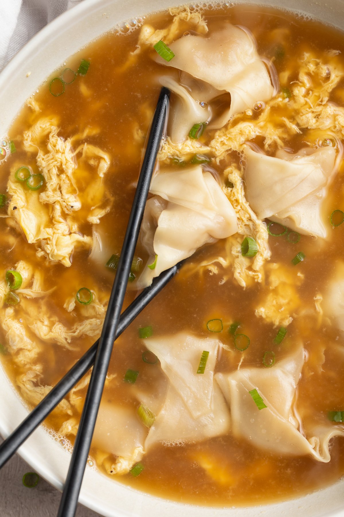 Overhead view of a bowl of wonton egg drop soup. 2 black chopsticks rest in the bowl of soup, and the right half of the bowl is out of the frame.