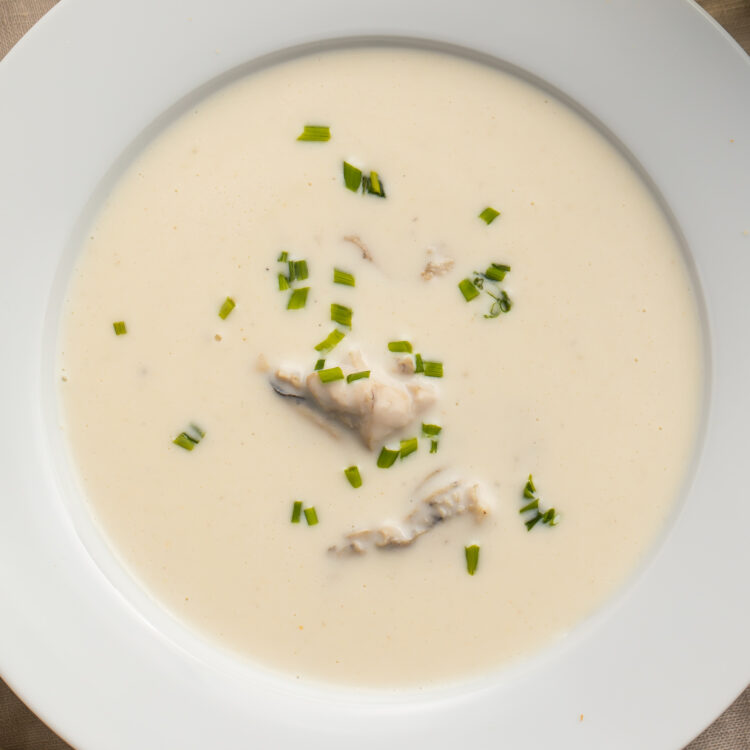 Close-up look at a soup bowl filled with pale, creamy oyster brie soup. Chunks of oyster rest in the center of the soup, and the top is dotted with fresh chives.