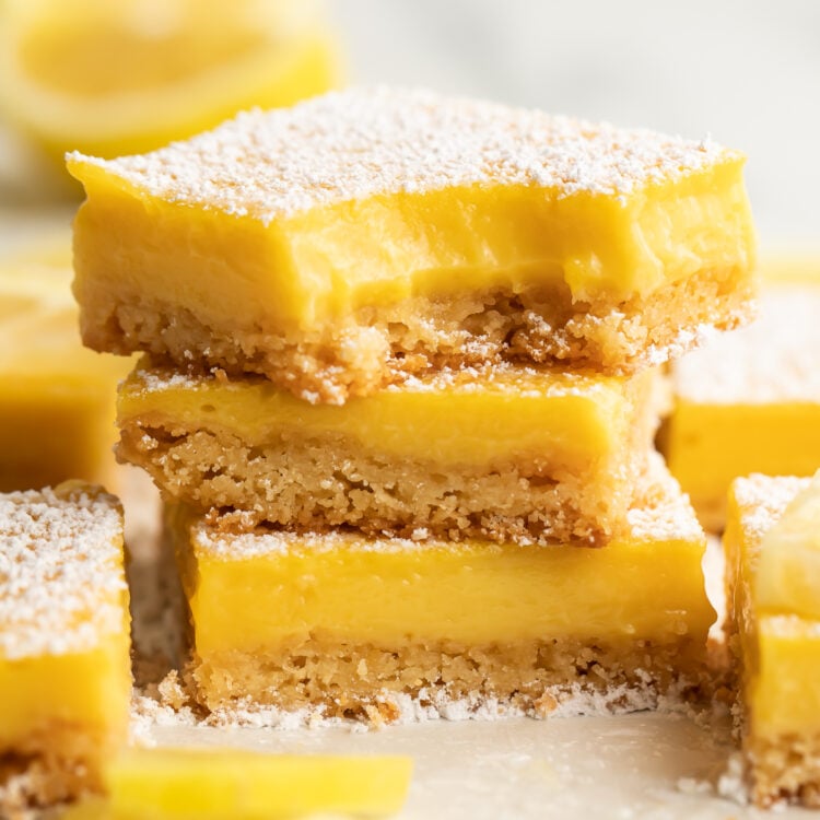 Three paleo lemon bars stacked on top of each other. A bite is missing from the corner of the lemon bar on top. Other lemon bars surround the stack, just out of focus.