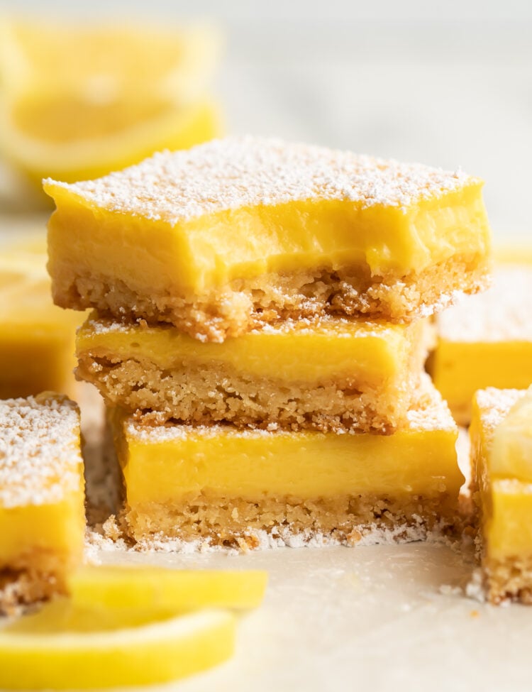 Three paleo lemon bars stacked on top of each other. A bite is missing from the corner of the lemon bar on top. Other lemon bars surround the stack, just out of focus.