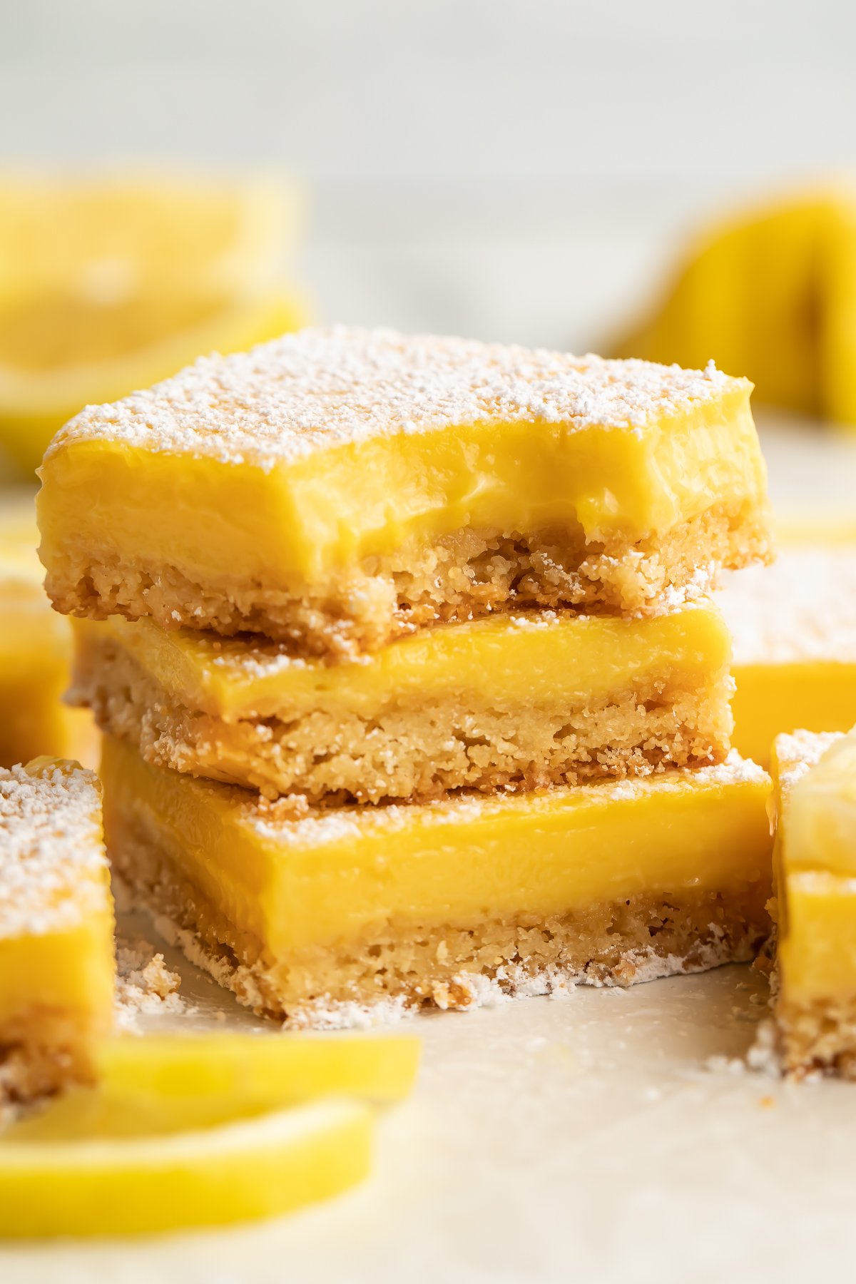 Three paleo lemon bars stacked on top of each other. A bite is missing from the corner of the lemon bar on top. Other lemon bars surround the stack, just out of focus.