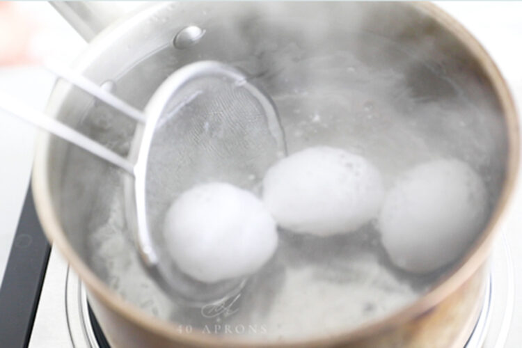 A handheld strainer lowering eggs into hot water.