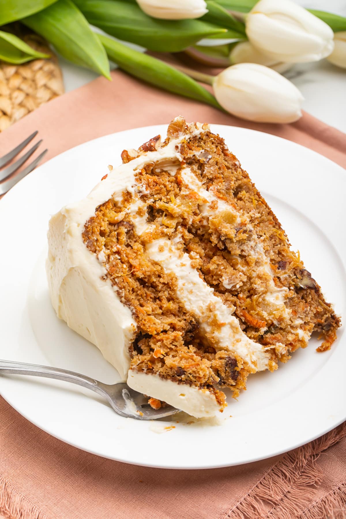 Slice of paleo carrot cake on a white plate resting on a light pink cloth napkin with a fork.