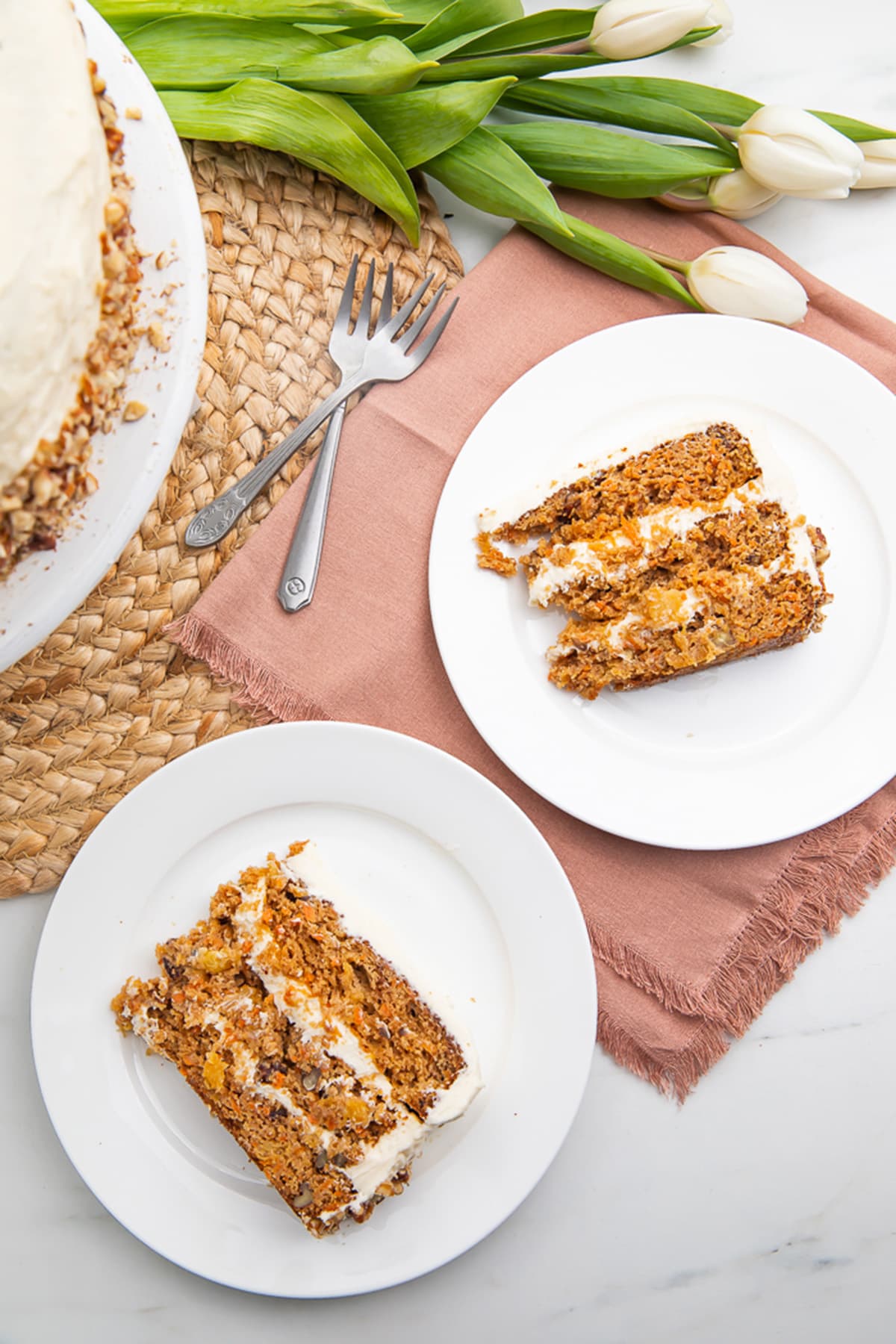 Top down view of 2 slices of gluten free carrot cake, each on a round white plate, sitting on light pink cloth napkins.