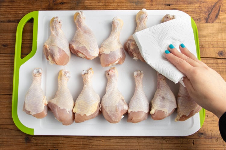 Chicken drumsticks on a cutting board. A woman's hand blots the chicken legs with a paper towel.