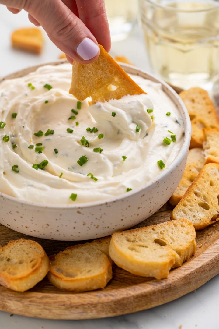 A white woman's hand using a crostini to scoop creamy boursin dip out of a white bowl.