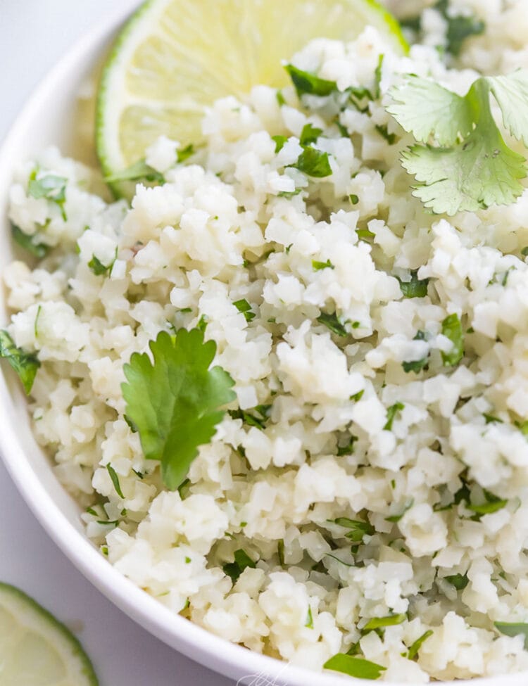 Cilantro lime cauliflower rice, garnished with a lime wedge, in a white bowl on a marble countertop.