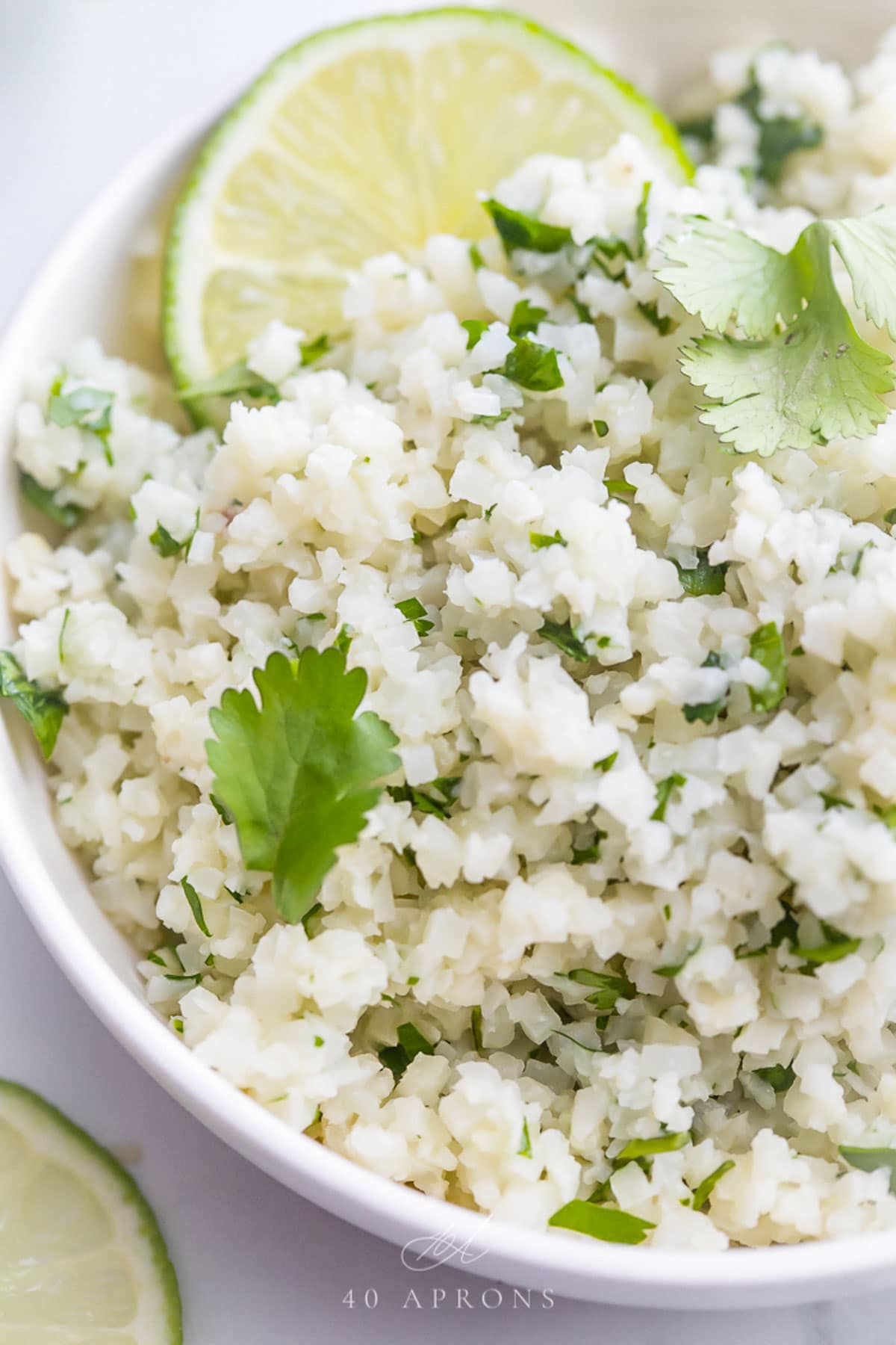 Cilantro lime cauliflower rice, garnished with a lime wedge, in a white bowl on a marble countertop.