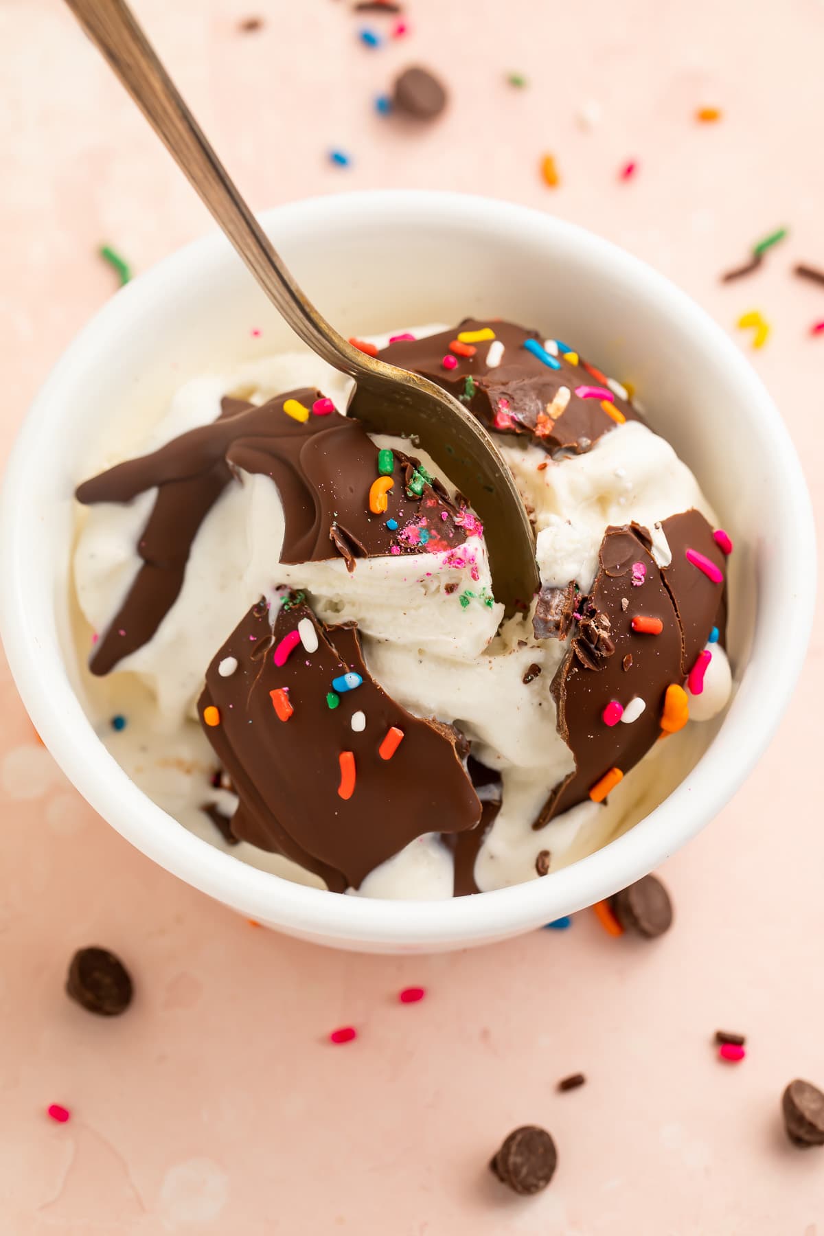 Broken chocolate magic shell topped with rainbow sprinkles over a bowl of vanilla ice cream.