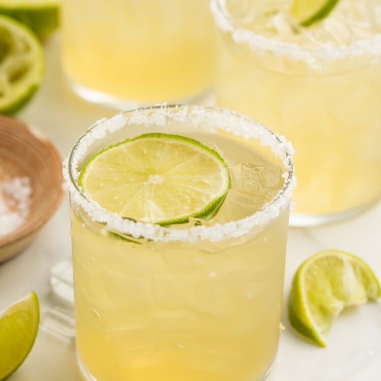 A salt-rimmed glass of virgin margarita garnished with a lime coin in front of 2 other glasses of margaritas.