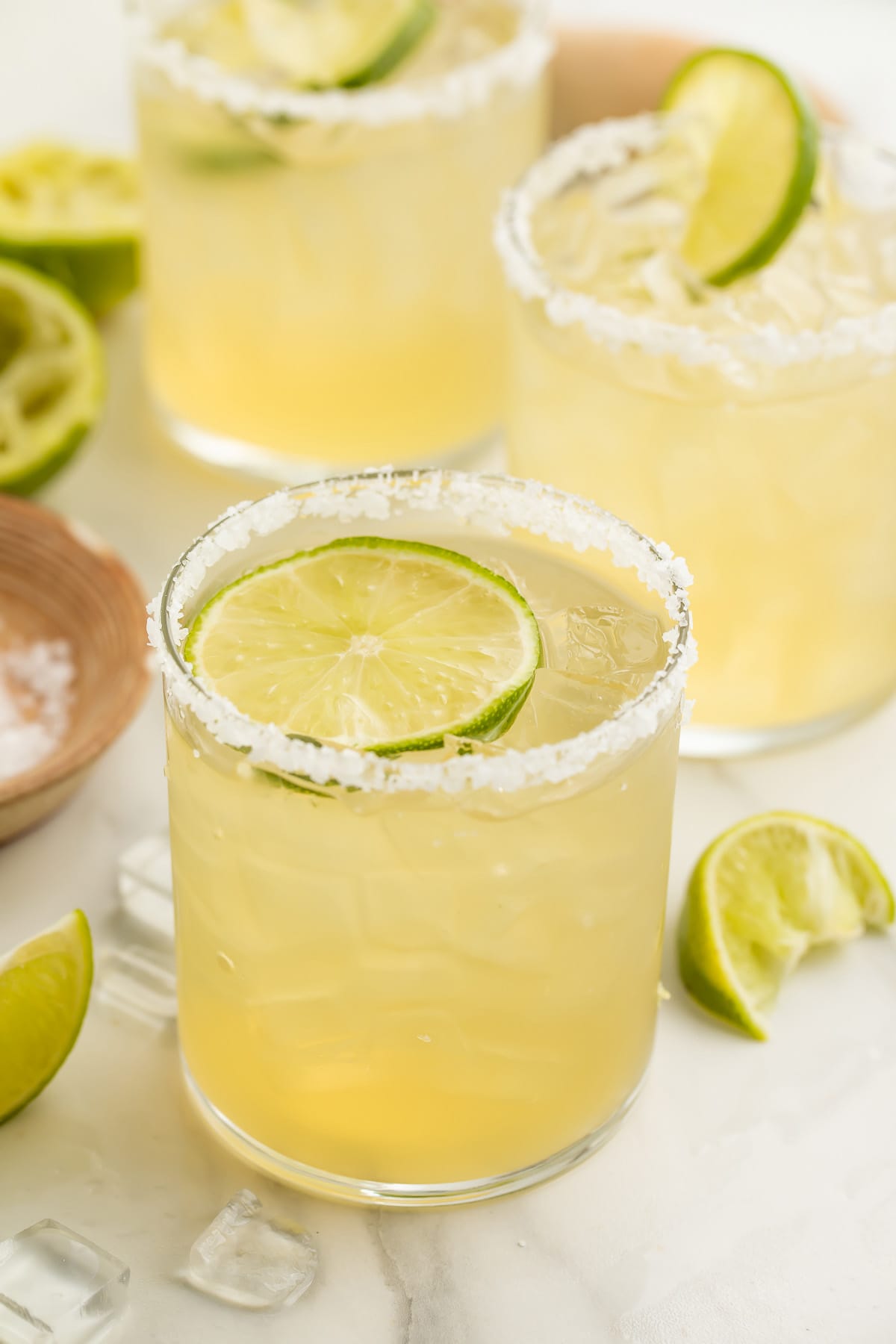 A salt-rimmed glass of virgin margarita garnished with a lime coin in front of 2 other glasses of margaritas.
