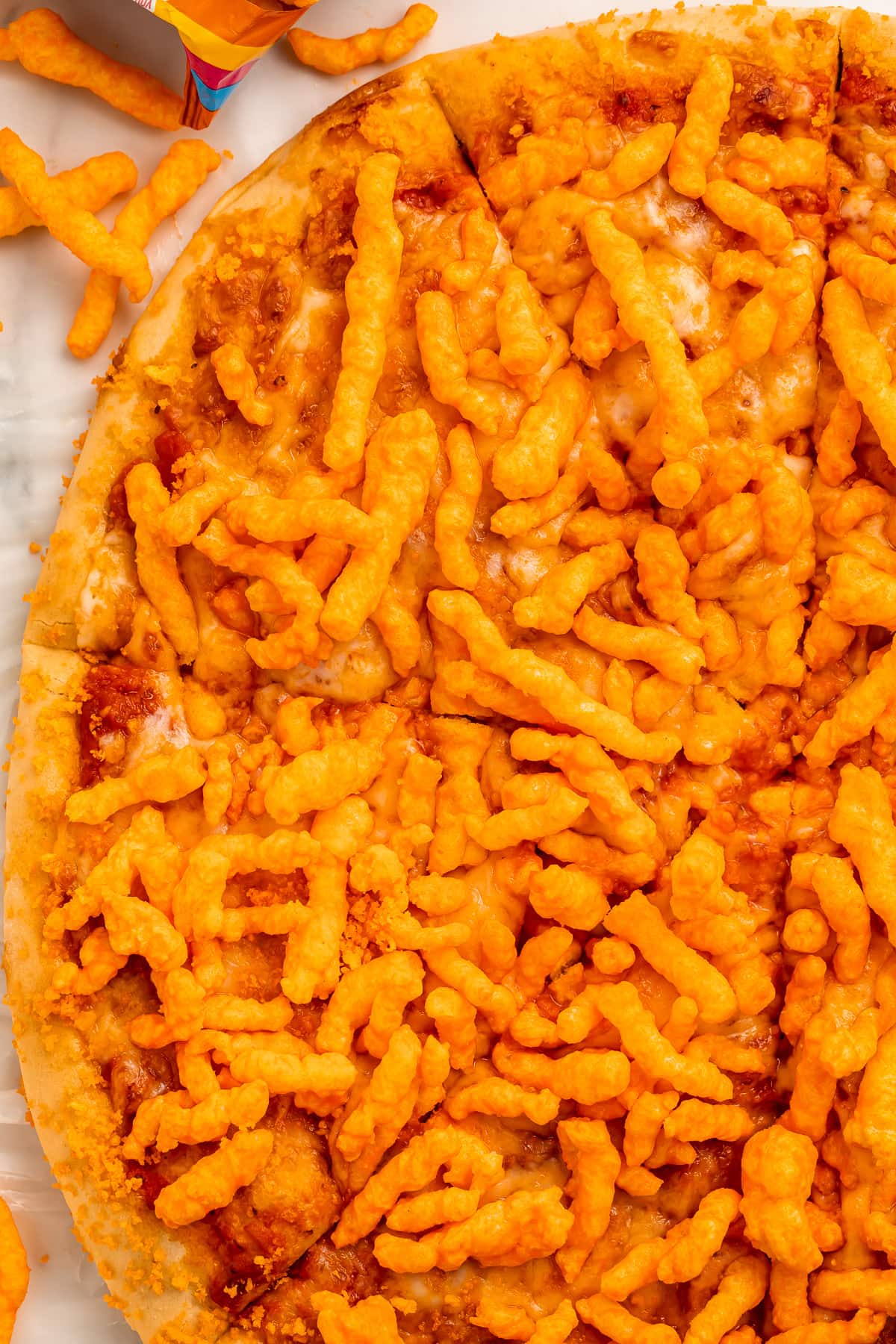 A large round cheese pizza topped with bright orange crunchy Cheetos, cut into triangle-shaped slices but still together as a whole pizza.