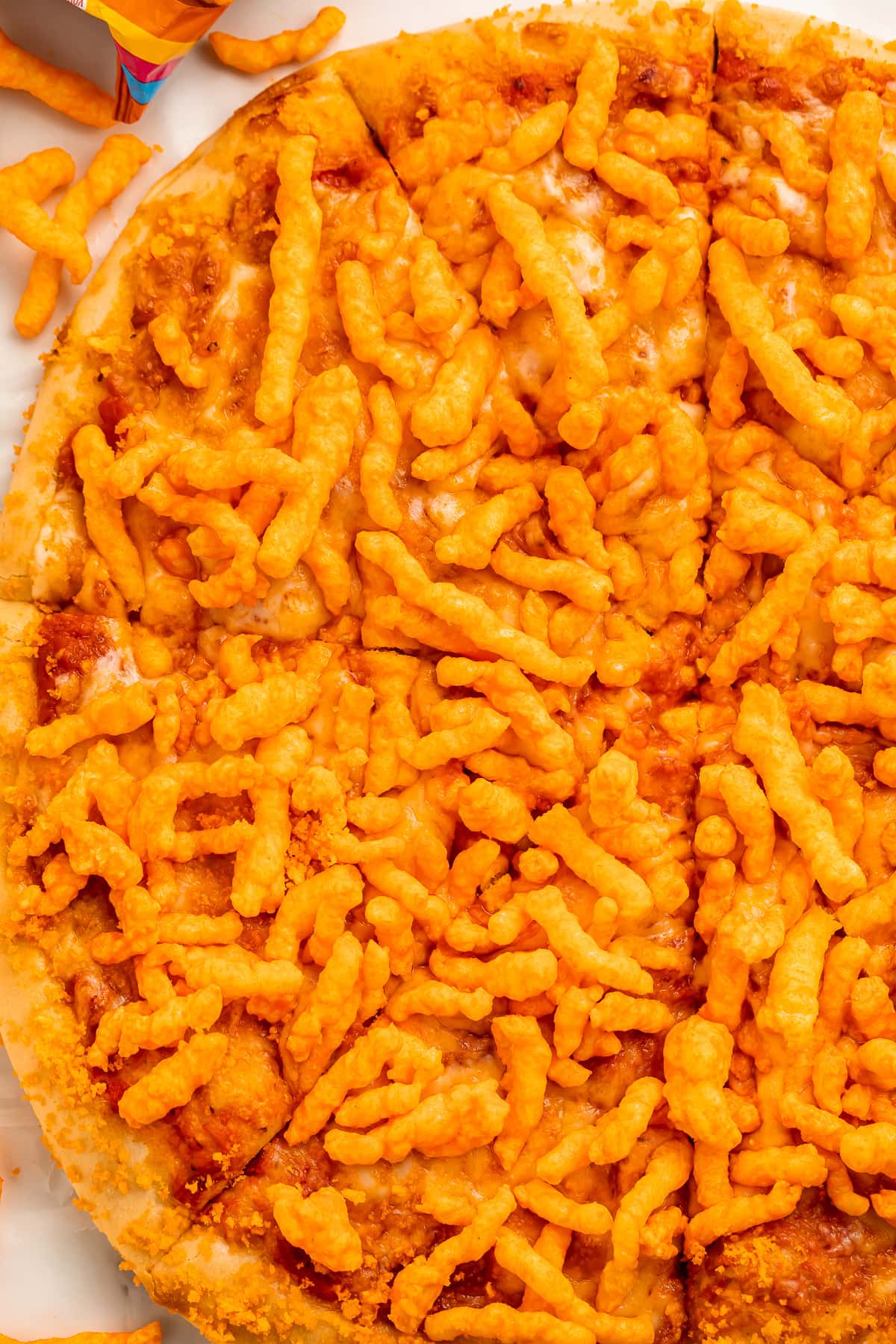 A large round cheese pizza topped with bright orange crunchy Cheetos, cut into triangle-shaped slices but still together as a whole pizza.