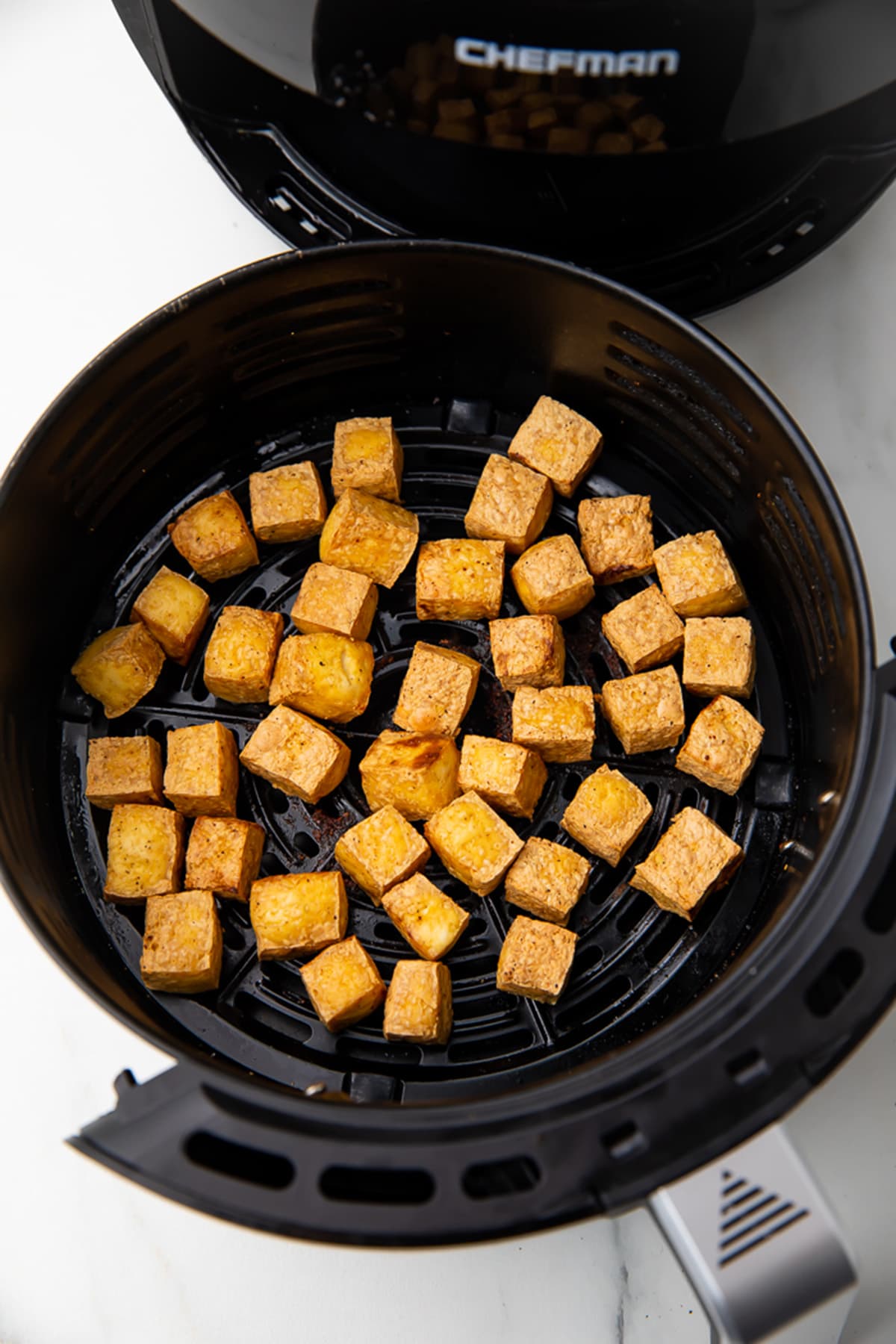 Air fried tofu in a round, black air fryer basket on a white countertop.