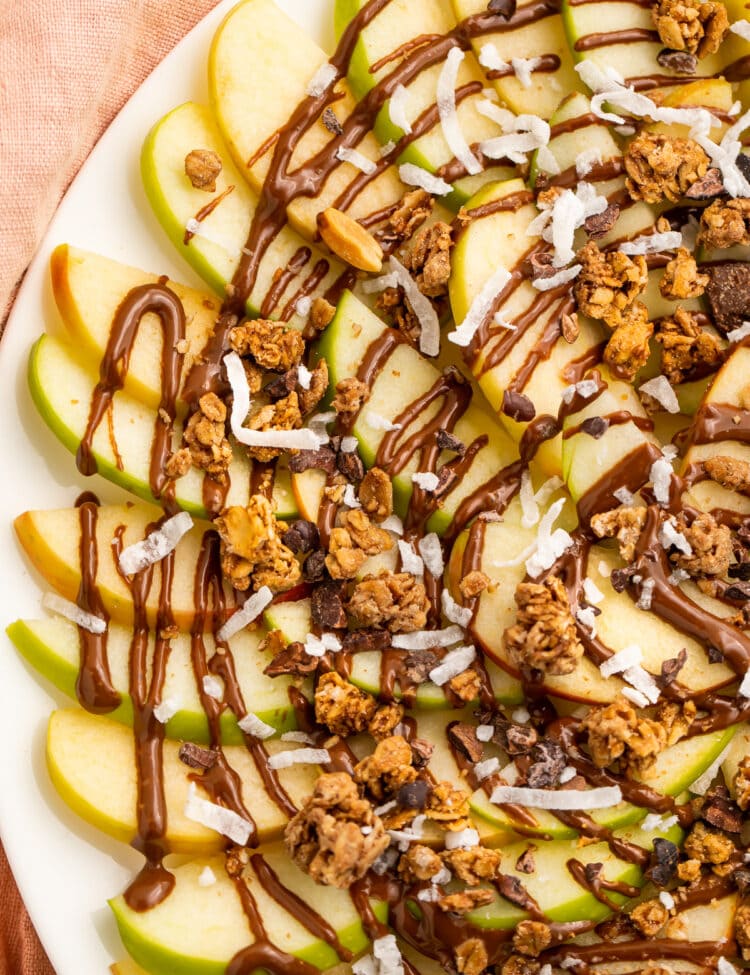 Overhead, top-down look at a plate of green and red apple slices topped with swirls of chocolate syrup and granola.